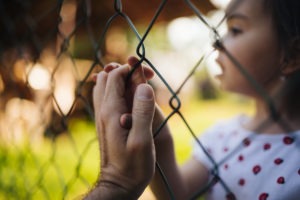 A man's hand reaches through a chain-link fence to touch his young daughter's hands.