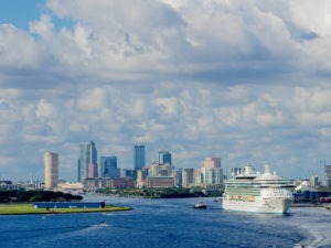 A white cruise ship sets sail away from Tampa Bay. The Tampa skyline stands in the background. It is a sunny day. The photo is taken from afar.