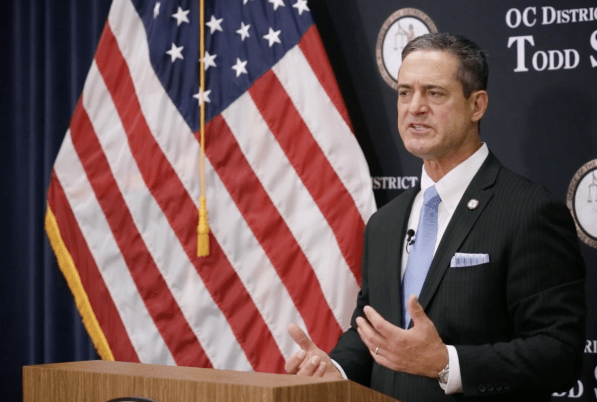 Orange County District Attorney Todd Spitzer stands in a suit behind a lectern and in front of an American flag.