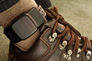 This photo shows a person's ankle with an electronic monitoring cuff attached. The person is wearing blue jeans and brown leather boots.