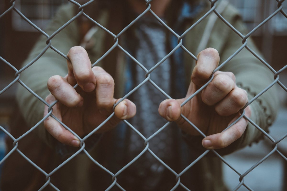 This photo shows a close-up of a person's hands gripping the inside of a prison fence, as if trying to get out.