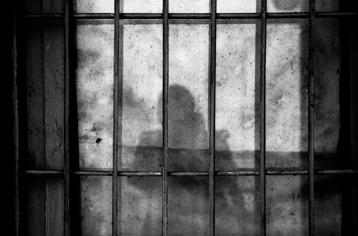 This photo shows the upper half of a person's shadow cast over metal jail-cell bars.