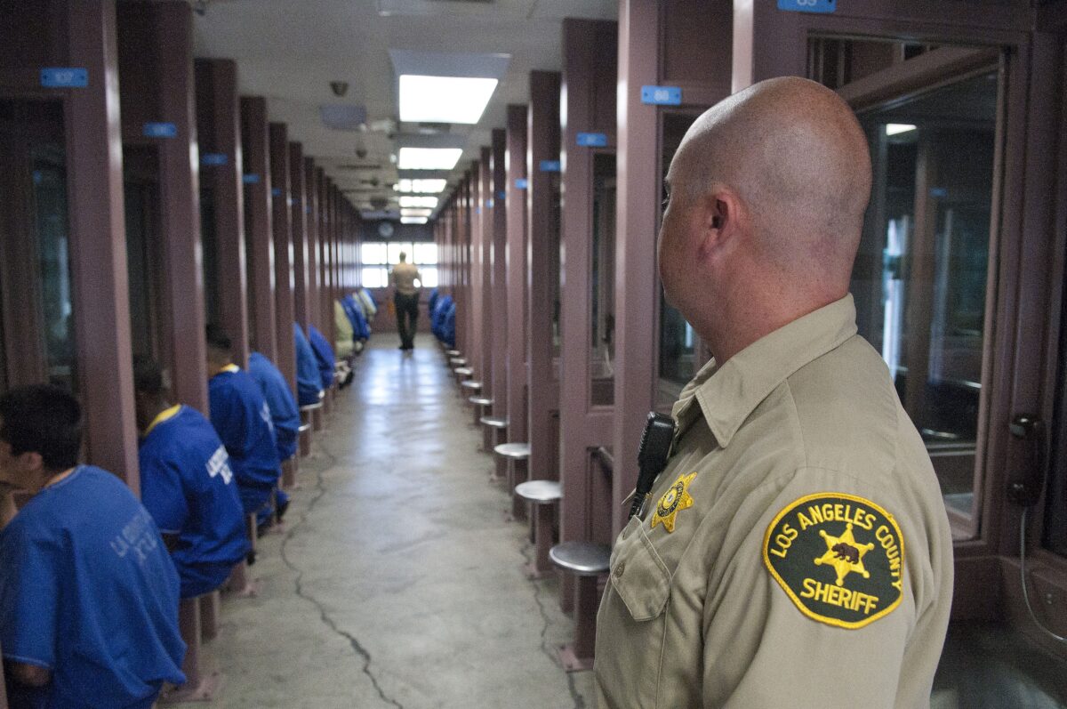This photo shows the back of a correctional guard's head as he stares down a long hallway. Prisoners sit in booths with their faces obscured, either visiting or using the phone.