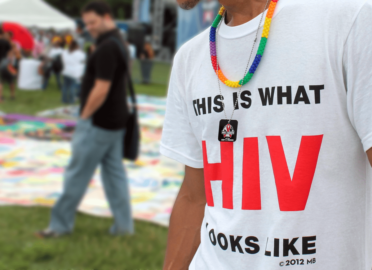This photo shows a close-up of a man wearing a white shirt that says "This is what HIV looks like." The picture is outdoors from the unveiling of a memorial AIDS quilt.