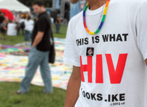 This photo shows a close-up of a man wearing a white shirt that says "This is what HIV looks like." The picture is outdoors from the unveiling of a memorial AIDS quilt.