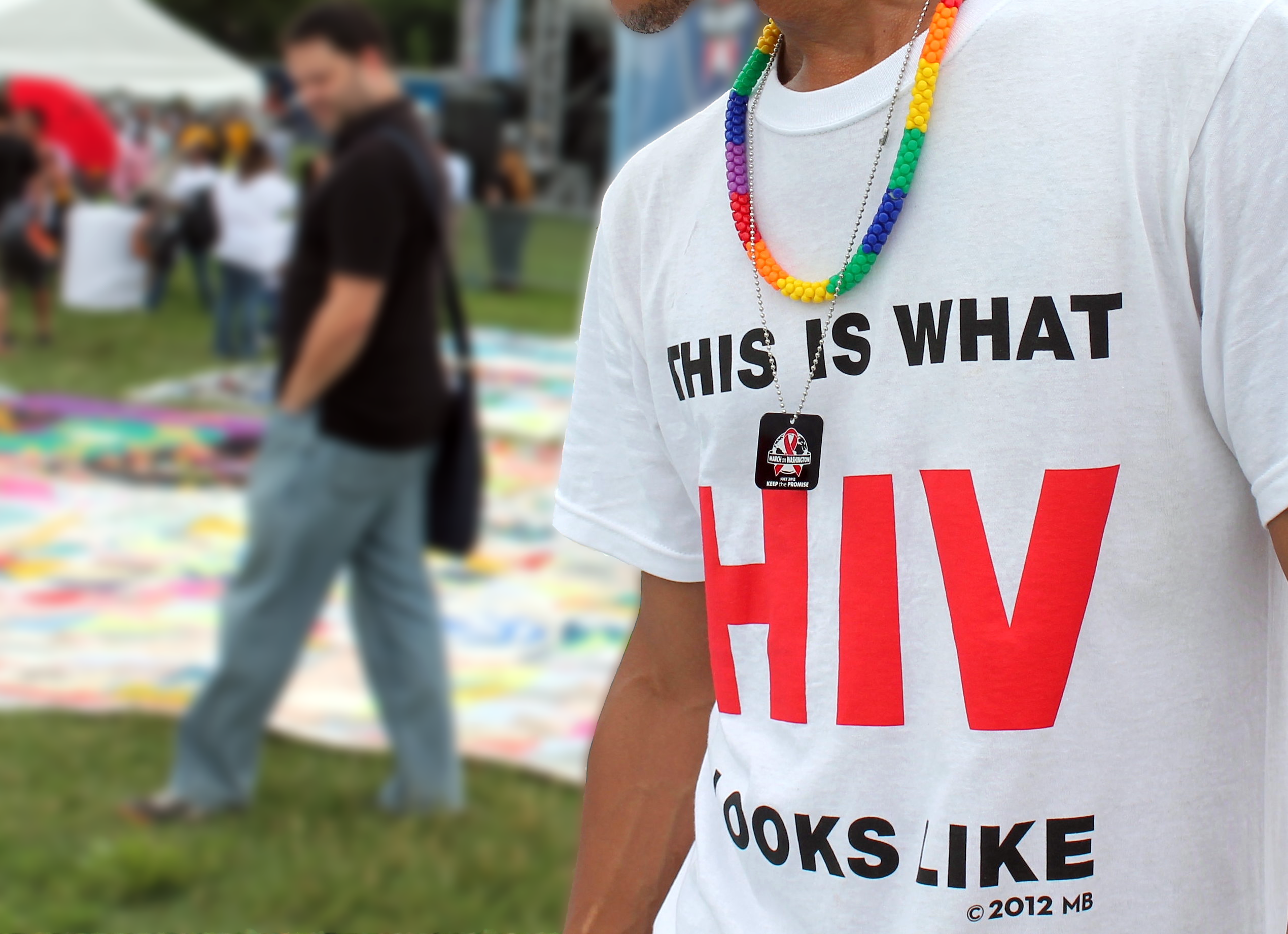 This photo shows a close-up of a man wearing a white shirt that says "This is what HIV looks like." The picture is outdoors from the unveiling of a memorial AIDS quilt.