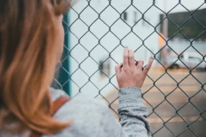 This photo shows a woman with red hair from behind, close up, over her right shoulder. She is clutching a fence and wearing a gray sweater. You cannot see her face.