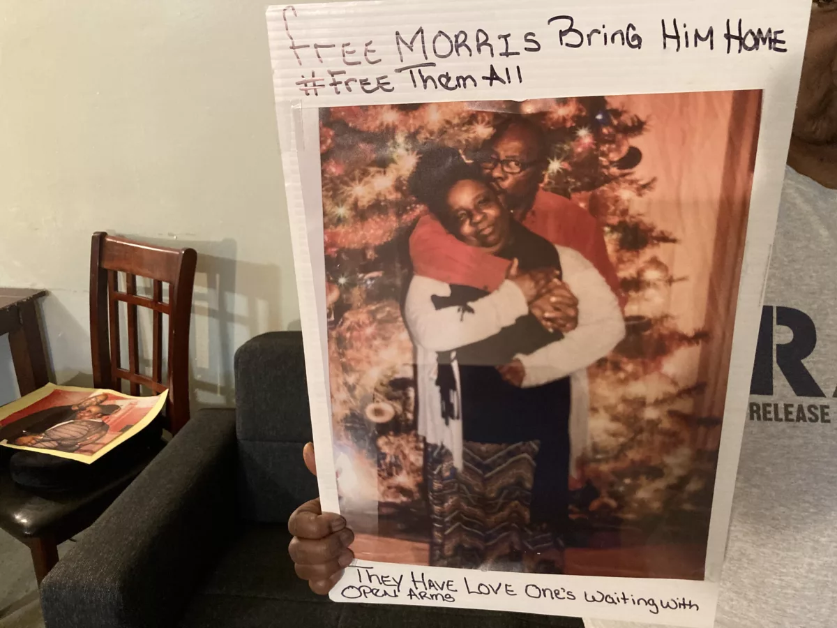 Theresa holding a sign that says "Free Morris! Bring Him Home" over a photo of her and Morris in front of a Christmas tree