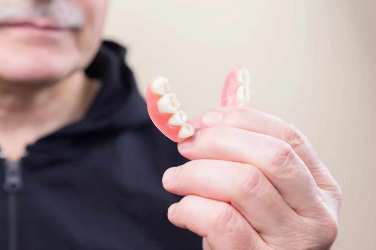This photo shows an elderly man holding a pair of dentures in front of his face. His full face is obscured, only his mouth can be seen. He is wearing a black sweater.
