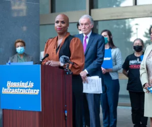 US Rep. Ayanna Pressley stands outdoors at a lectern. The lectern has a sign on the front that says "housing is a human right." She is wearing a rust-colored shirt. U.S. Senator Ed Markey stands behind her.