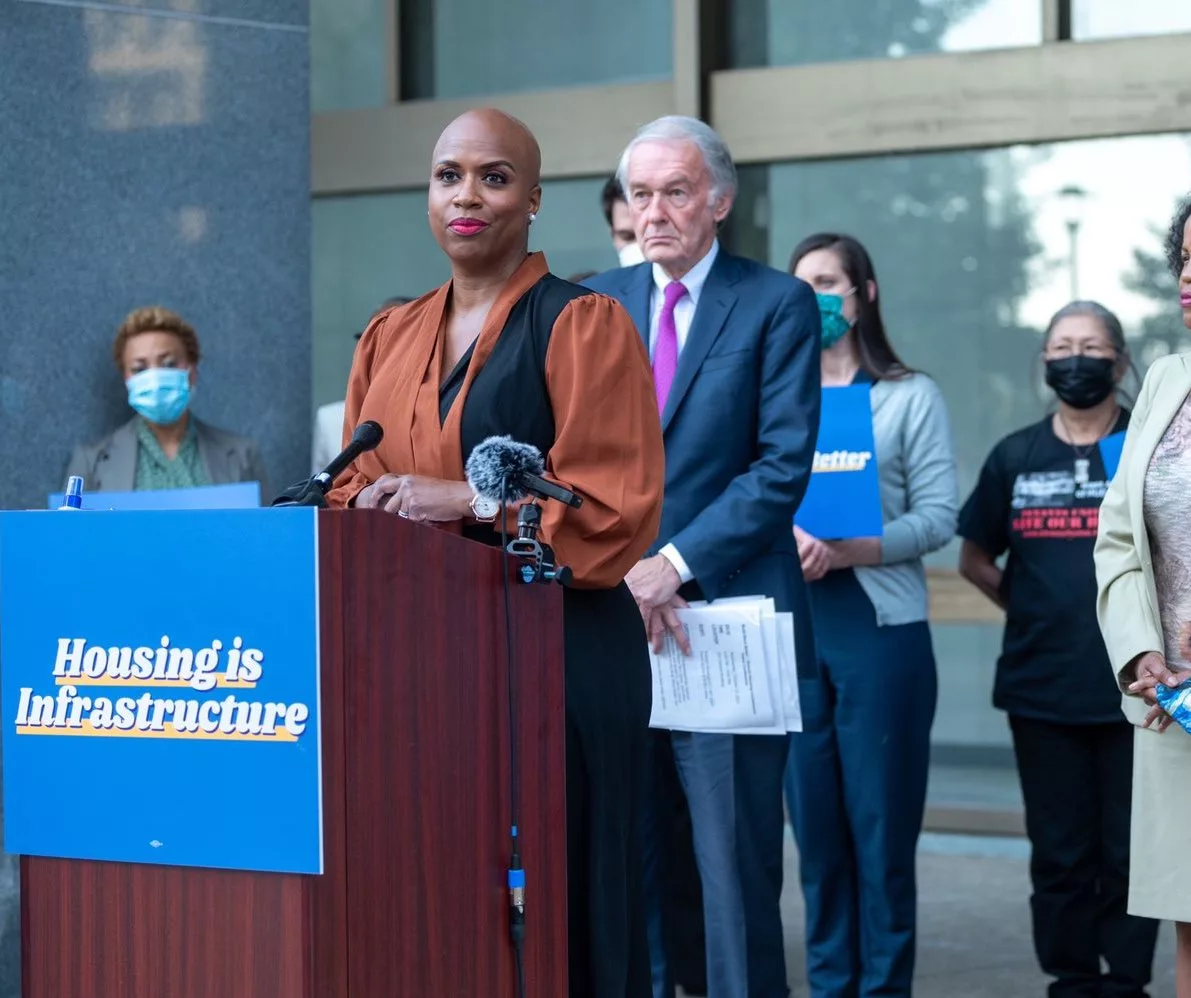 US Rep. Ayanna Pressley stands outdoors at a lectern. The lectern has a sign on the front that says "housing is a human right." She is wearing a rust-colored shirt. U.S. Senator Ed Markey stands behind her.