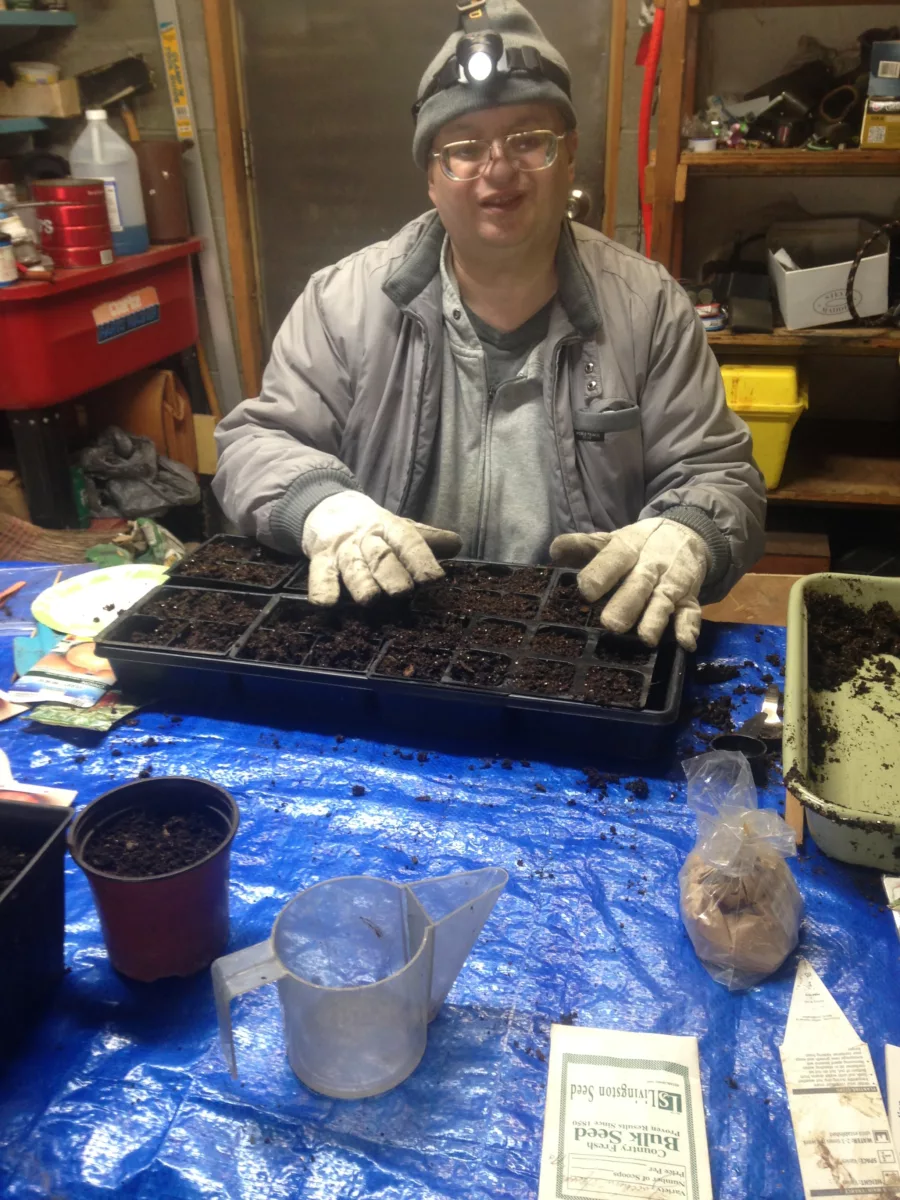 Anthony Talotta sits at a table in front of a bunch of soil. He is gardening. He is smiling.