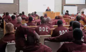 This photo shows people imprisoned in Pennsylvania wearing jumpsuits while listening to someone speak at a podium.