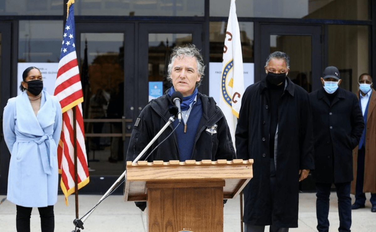 Cook County Sheriff Thomas J. Dart stands at a lectern speaking outdoors.