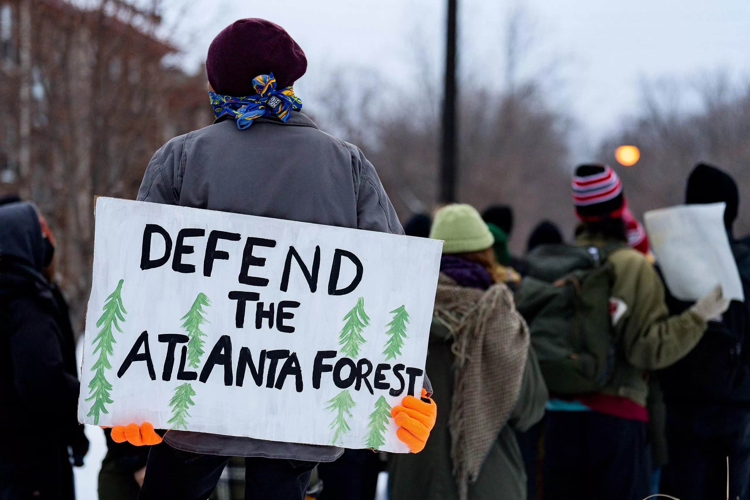 A fully masked, anonymous person in cold weather holds a sign that says "defend the Atlanta forest" in an anti Cop City protest.