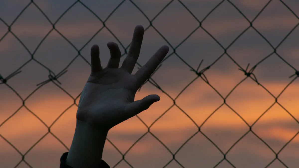 This photo shows the silhouette of a person's hand against a cyclone fence.