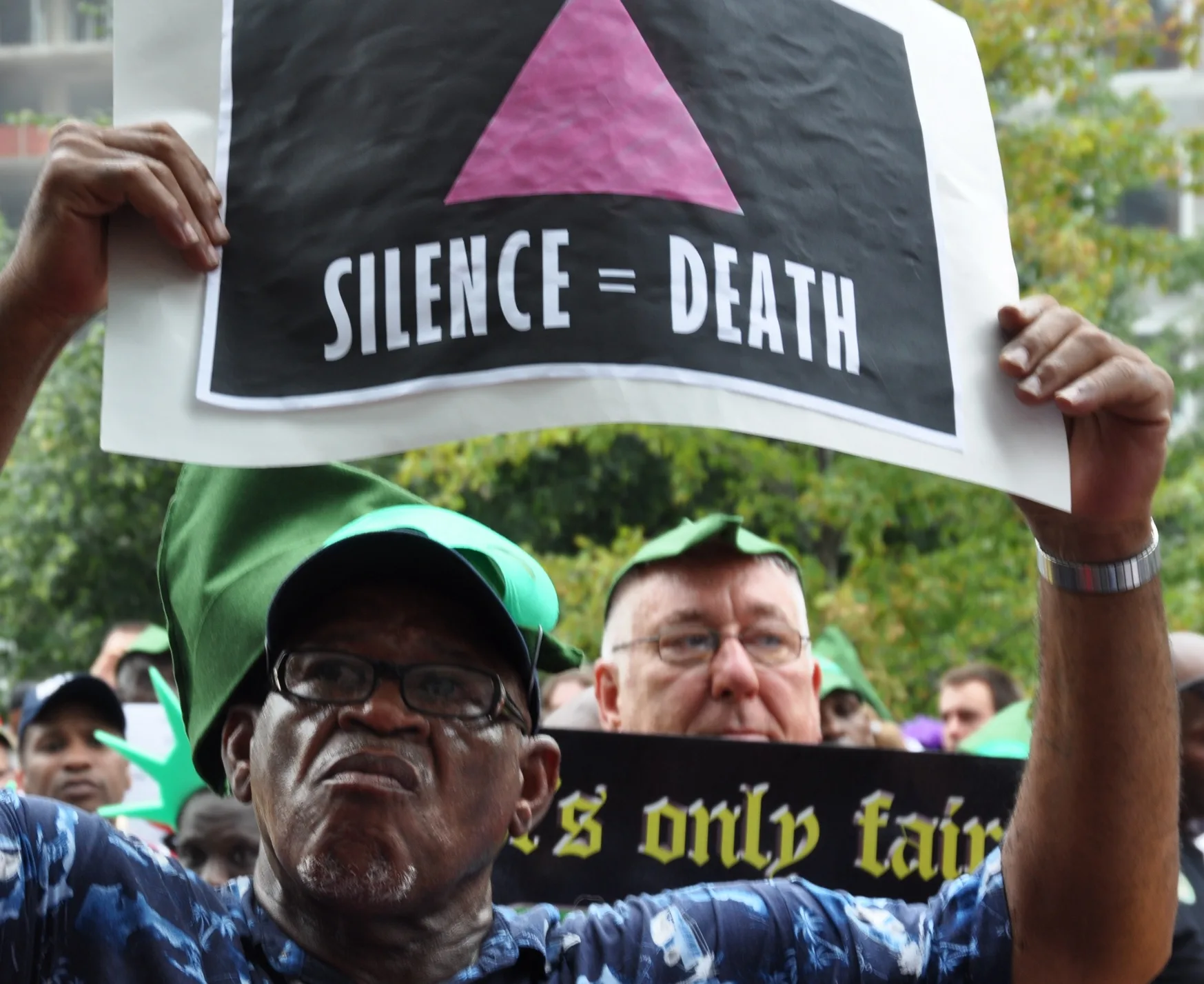 This photo shows a man outdoors at a protest holding an Act Up sign that states "Silence = Death." The sign is black with white lettering and a pink triangle.