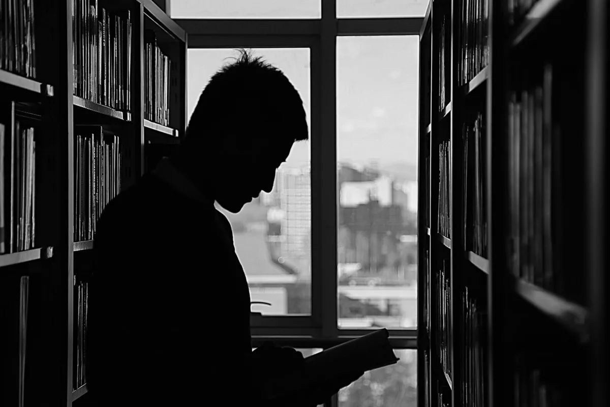 This photo shows a silhouette of a man in a library. He stands by a window reading a book.