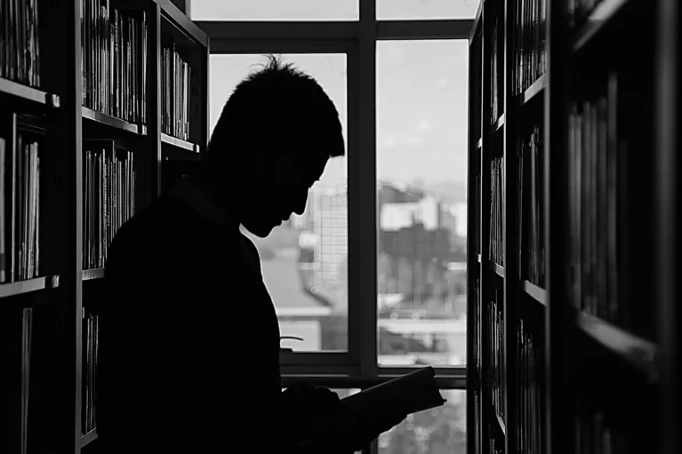 This photo shows a silhouette of a man in a library. He stands by a window reading a book.