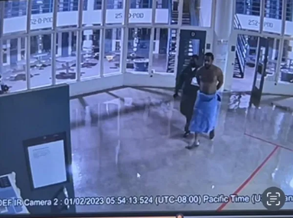 This photo is a screenshot of surveillance footage of Stanley Wilson. Wilson is led, handcuffed and wrapped in a towel, through a jail by an officer.