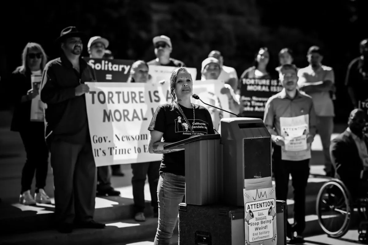 Jessica Solis delivers a speech about solitary confinement outside the State Capitol. She stands at a lectern as protesters hold signs behind her.