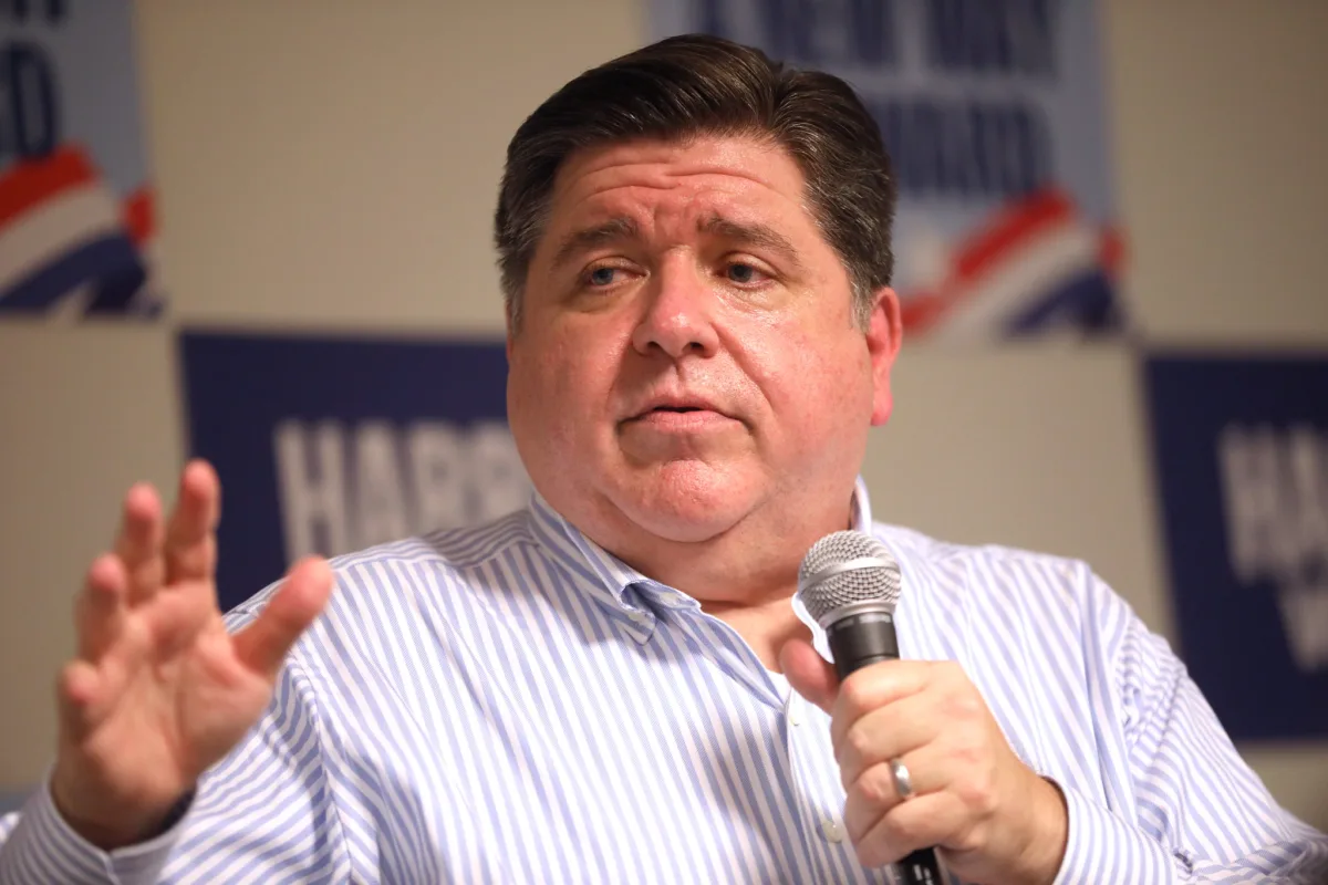 This photo shows a close-up of Illinois Gov. JB Pritzker giving a speech. He holds a microphone and is wearing a seersucker striped dress shirt.