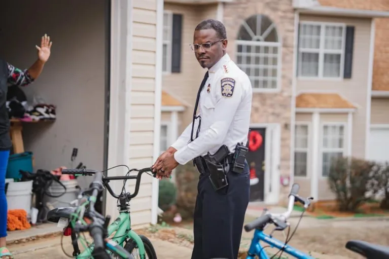 Clayton County Sheriff Levon Allen poses for a staged photograph holding a child's bicycle outdoors. He is wearing his uniform and making a serious expression with his face.