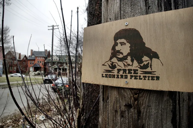 A sign on a tree with a stenciled image of Peltier and the words "Free Leonard Peltier"