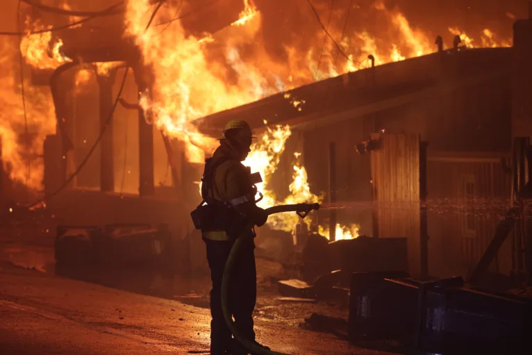 This photo shows a California firefighter spraying a burning house with a hose at night during the 2025 Palisades Fire.