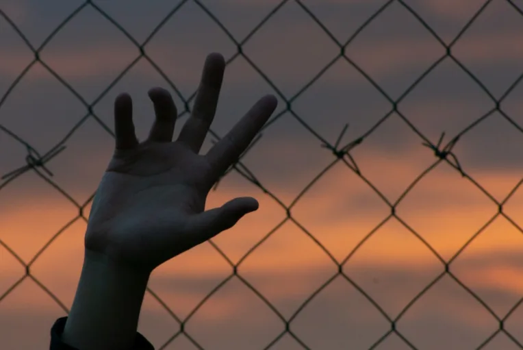 This photo shows a child's hand reaching toward the sky, but it is behind a chain-link fence.