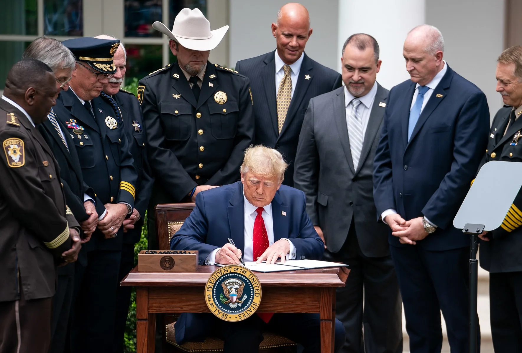 This photo shows President Donald Trump in 2020 signing an executive order outdoors. He is seated at a table surrounded by police chiefs.